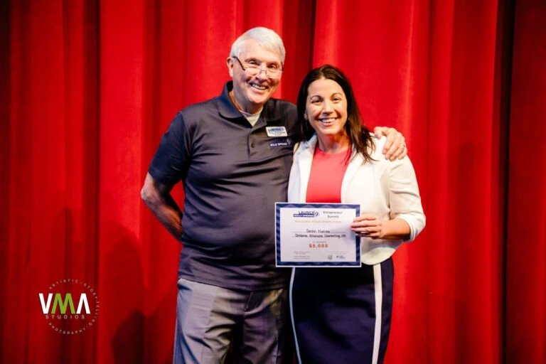 Two smiling people on stage, one holding a check, in front of red curtains.