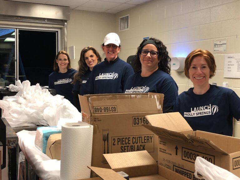 Five women in matching blue shirts smiling in a room with boxes and packing supplies.