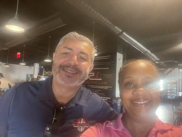 Two smiling people in a gym or fitness center, with workout equipment in the background.