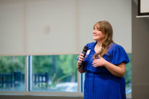 Woman in blue dress speaking into a microphone during a presentation.