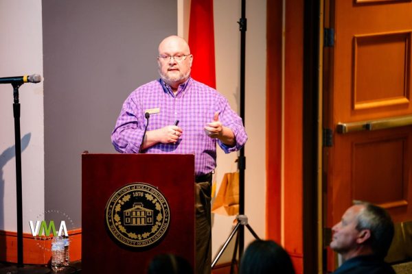 Bald man in purple checkered shirt speaking at a podium with a university seal.