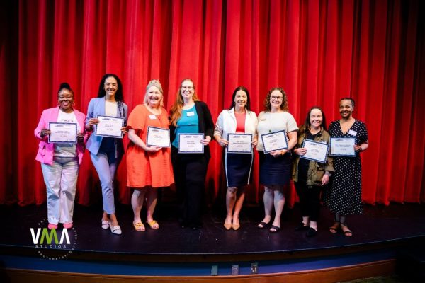 Group of eight women on stage holding certificates in front of red curtain.