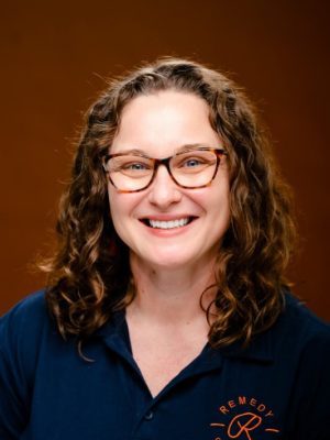 Smiling woman with curly hair and glasses wearing a navy polo shirt.