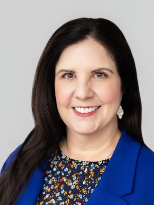 Smiling woman with long dark hair wearing a blue blazer and floral top against a light gray backgrou.