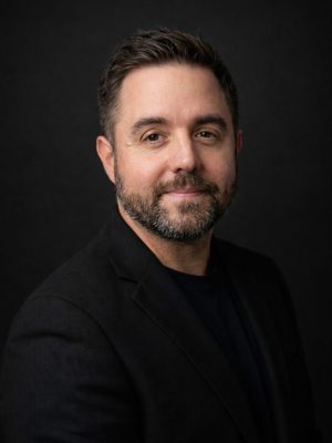 A man with dark hair and a beard, wearing a black blazer, smiling softly against a dark background.