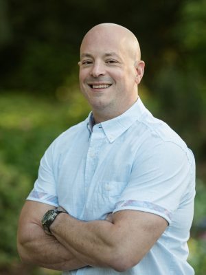 Smiling bald man in light blue shirt standing outdoors with arms crossed.