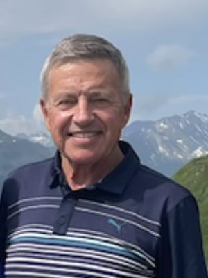 A man with gray hair smiling outdoors with mountains in the background.