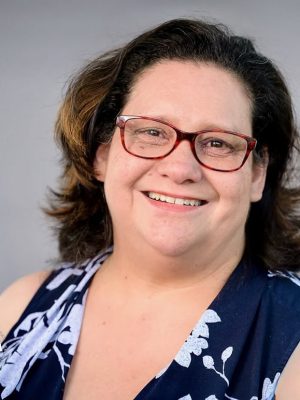 Woman with glasses smiling, wearing a blue and white floral top.