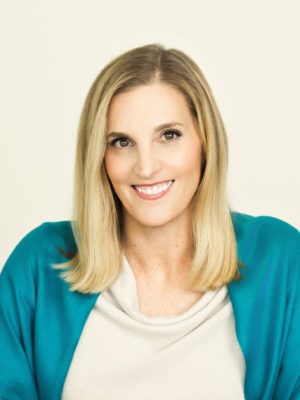 A woman with blonde hair smiling at the camera, wearing a white top and a teal cardigan.
