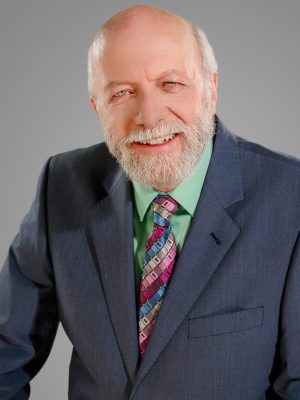A smiling older man with a beard wearing a dark suit, green shirt, and colorful tie against a gray b.